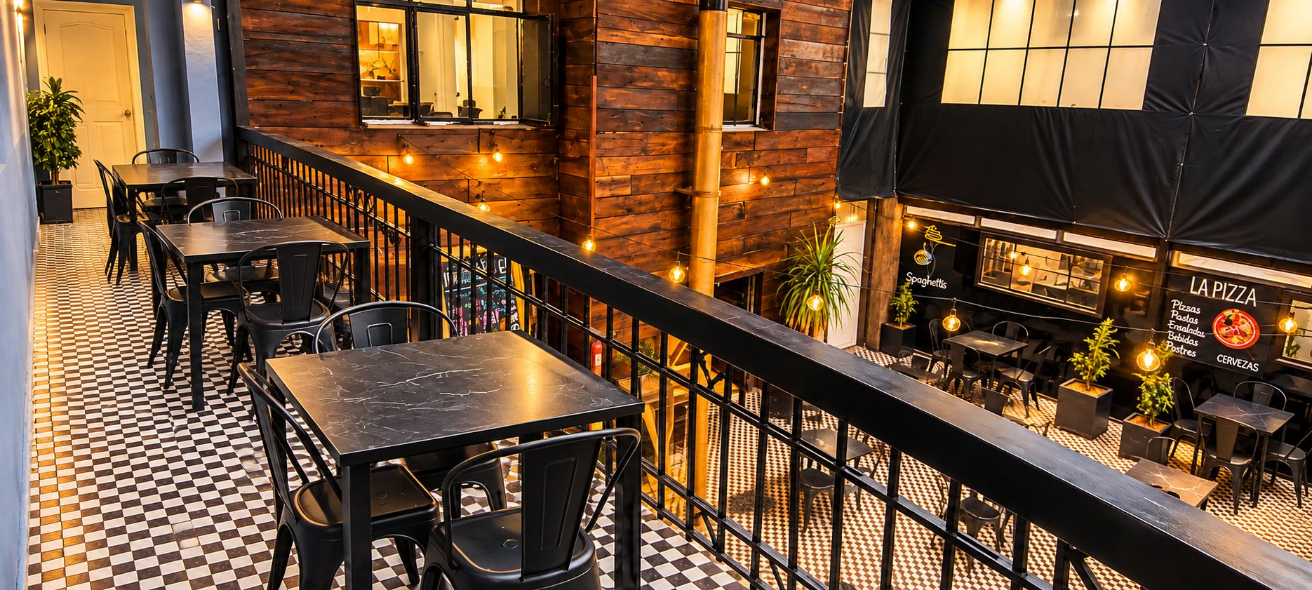 An outdoor dining area with checkered flooring, metal tables, and chairs, overlooking a restaurant with a sign for 'LA PIZZA' and 'SPAGHETTI'.