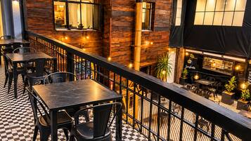 An outdoor dining area with checkered flooring, metal tables, and chairs, overlooking a restaurant with a sign for 'LA PIZZA' and 'SPAGHETTI'.