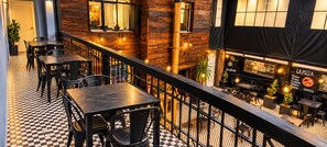 Un área de comedor al aire libre con piso a cuadros, mesas y sillas de metal, con vista a un restaurante que muestra un letrero que dice 'LA PIZZA' y 'SPAGHETTI'.