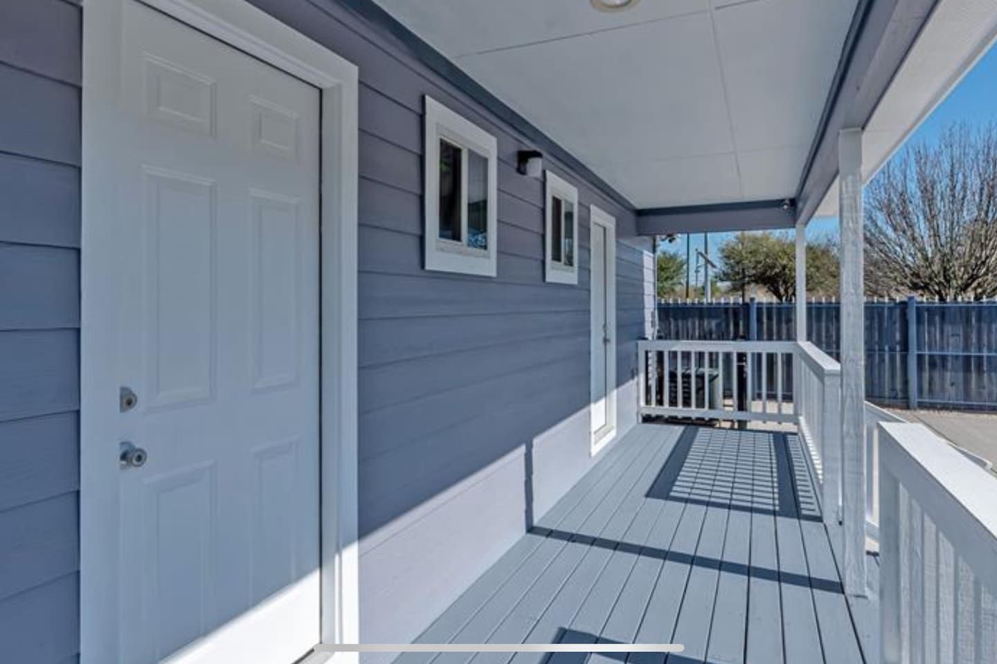 A covered porch with a white door and windows, a blue fence, and a clear sky.