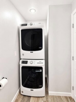 A stacked washer and dryer unit in a corner of a room with white walls and a wooden floor. - Gated Loft near NRG and Texas Med Center (Houston)