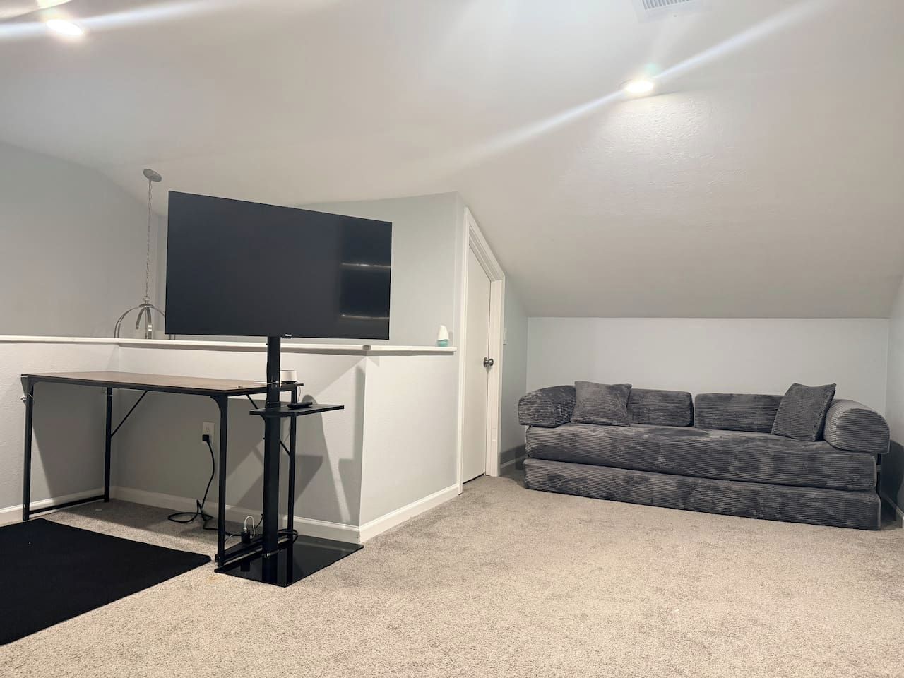 A room with a desk, a flat-screen TV mounted on the wall, a grey sofa, and a black mat on the floor.