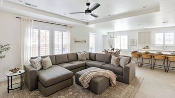 A modern living room with a sectional sofa, a dining area with a kitchen in the background, and a ceiling fan.