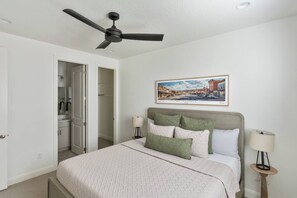 A bedroom with a bed, a ceiling fan, a painting of a cityscape, and a nightstand with a lamp.