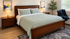 A bedroom with a wooden bed, two bedside tables, a nightstand, a floor lamp, a potted plant, and a window with blinds.