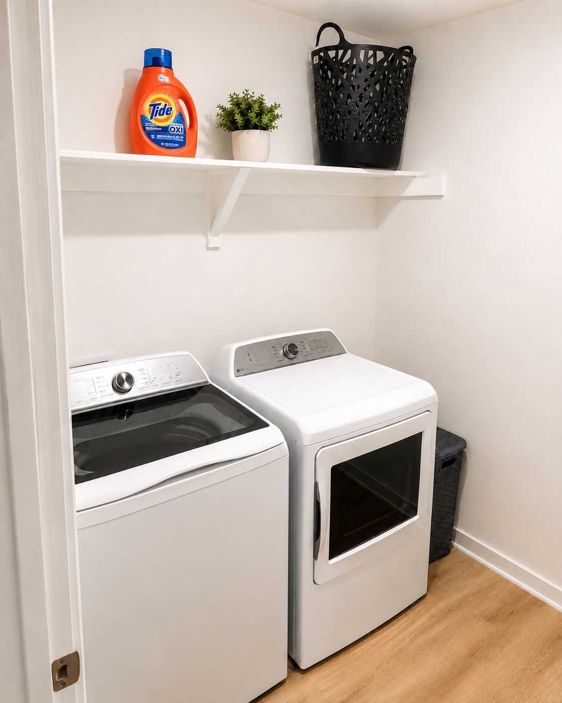 Un espace buanderie comprenant deux machines à laver à chargement frontal, une bouteille de lessive, une petite plante et un panier noir posé sur une étagère.