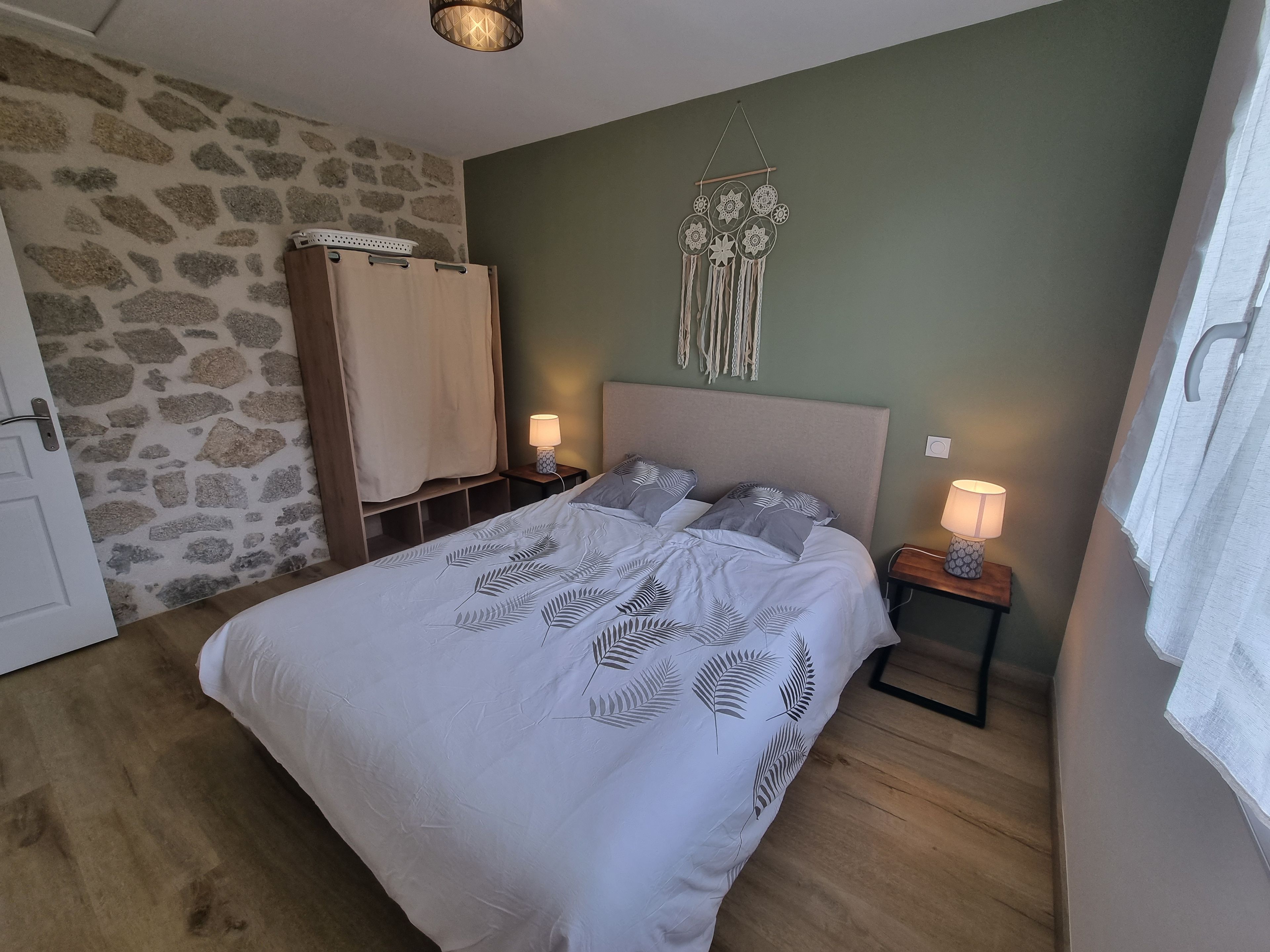 Un dormitorio con una cama, dos mesitas de noche con lámparas, un atrapasueños colgado en la pared y una pared con diseño de piedra.