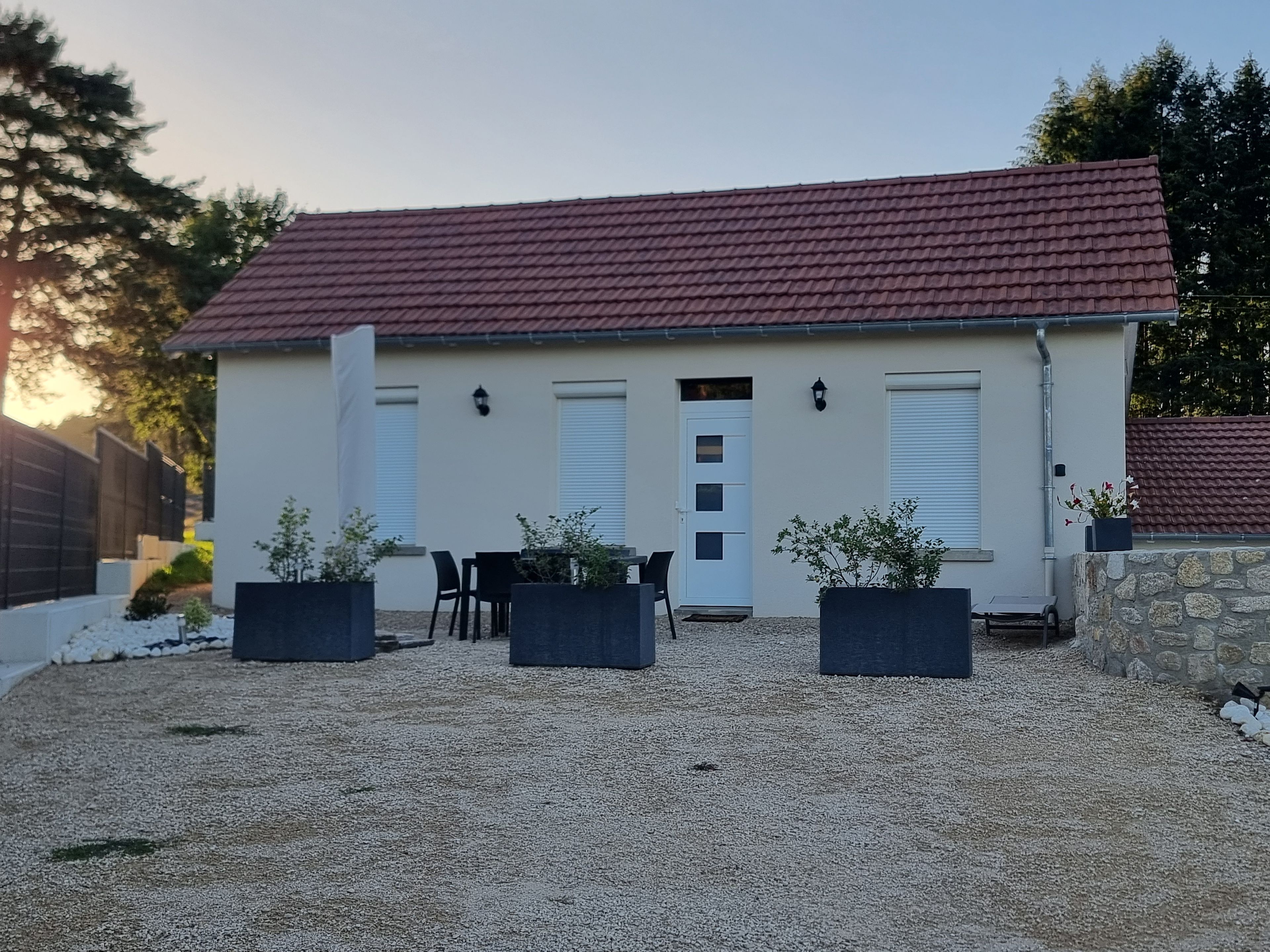 A single-story building with a red-tiled roof, a white door, and two windows with shutters. There are three black planters with plants in front of the building.