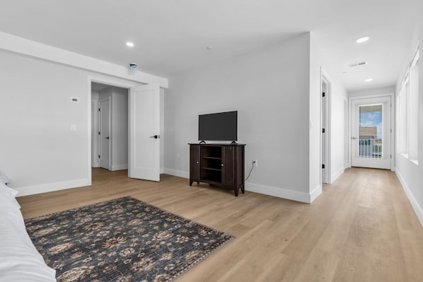 A spacious room with hardwood flooring, a dark patterned rug, a flat-screen TV on a wooden stand, and white walls.