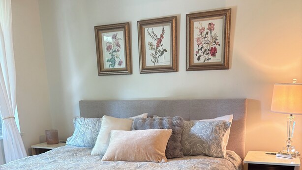 Ein ordentlich bezogenes Bett mit einem gemusterten Überwurf, ein Nachttisch mit einer Lampe und drei gerahmte Pflanzenbilder an der Wand.