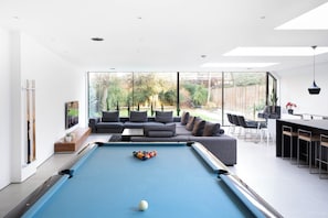 Een moderne woonkamer met een pooltafel, grijze banken en een groot raam met uitzicht op een tuin.