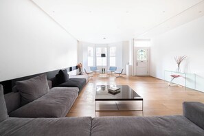 A modern living room with a grey sofa, a glass coffee table, and a dining area with a glass table and chairs. - Large Modern London Townhouse with Cinema, Gym, Garden, Pool table, Large spaces (London)