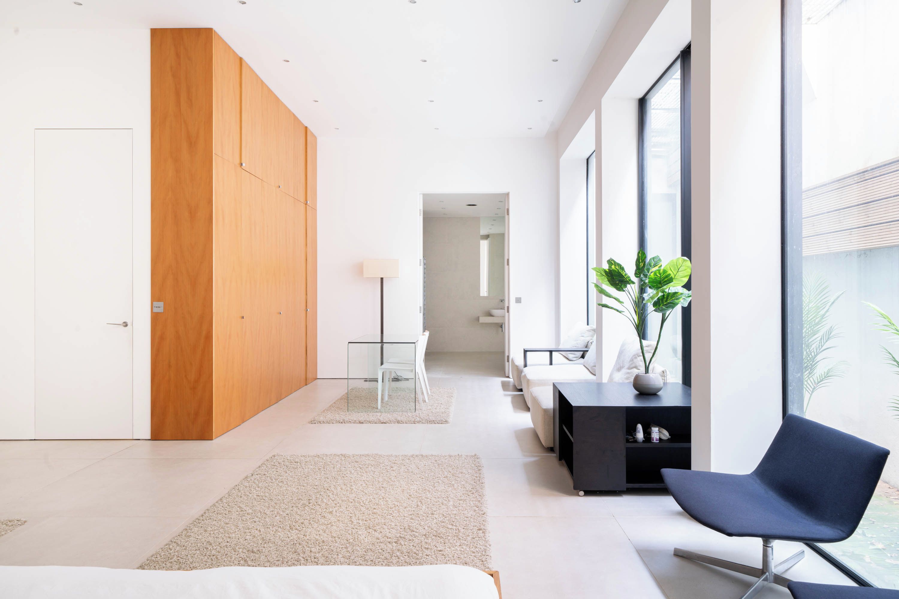 Una habitación de hotel moderna con una cama grande, un armario de madera, un escritorio con silla, un sillón negro y ventanas amplias que ofrecen una vista.