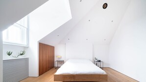 Ein modernes Schlafzimmer mit schrägen Wänden, einem Holzbettgestell, einem Nachttisch mit Lampe und einer kleinen Pflanze auf dem Fensterbrett.