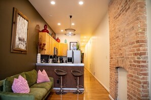 A compact living space with a kitchenette, a green sofa with patterned cushions, and a brick wall. - Eclectic Comfort: The Kitt – 2BR/2BA w/ Balcony Near MICA & JHU (Baltimore)