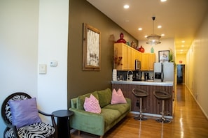 A compact living space with a kitchenette, a green sofa with patterned cushions, a small round table, and a bar stool. - Eclectic Comfort: The Kitt – 2BR/2BA w/ Balcony Near MICA & JHU (Baltimore)