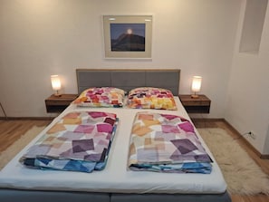 Un llit doble amb un edredó de disseny geomètric i colors, dues taules de nit amb làmpades, un quadre amb marc a la paret, i una catifa esponjosa al terra.