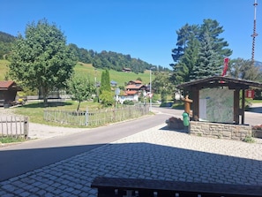A paved area with a wooden bench, a small kiosk with a map, and a road leading to houses in a hilly area.