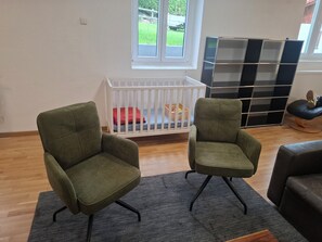 A room with two green armchairs, a white crib, a grey rug, and a black and white shelving unit.