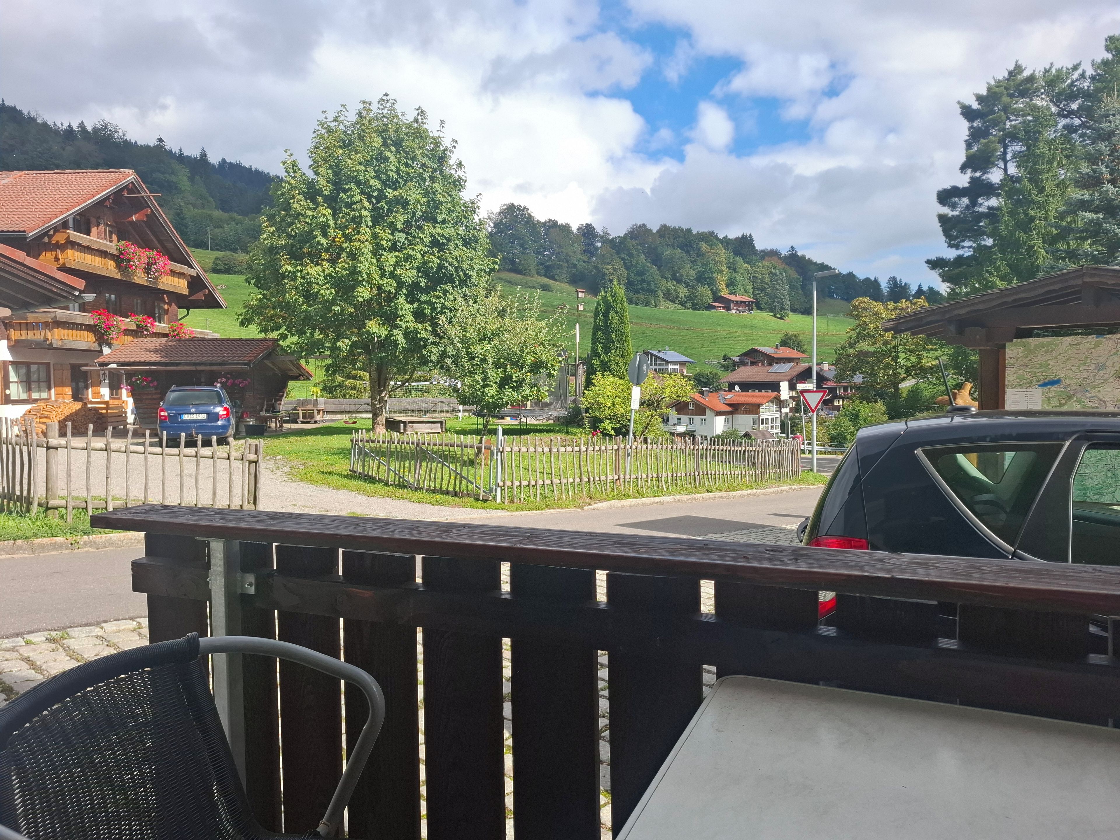 A wooden deck with a chair, a car, and a view of a village with houses and a mountain in the background.