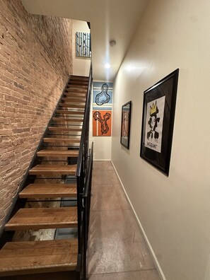 A staircase with wooden steps and metal railings, flanked by brick walls and artwork. - Colorful & Quirky 1-BR Loft w/Private Deck | MICA, JH & UMMC (Baltimore)