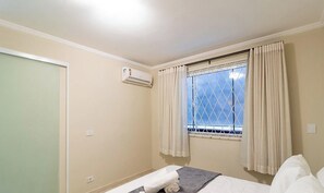 A room with a bed, an air conditioning unit, a window with curtains, and a view of the sky.