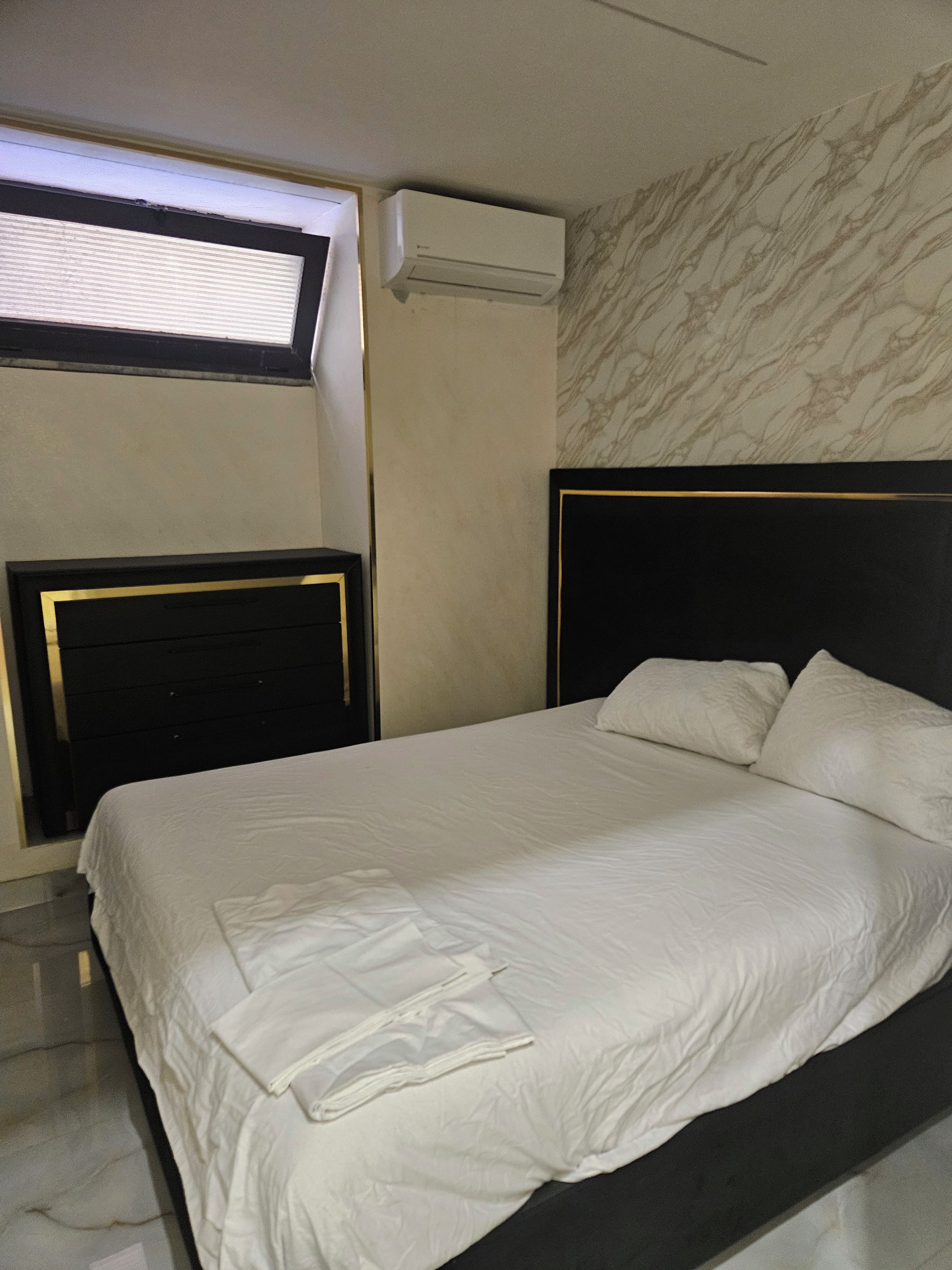 A hotel room with a bed, a window with blinds, an air conditioning unit, and a marble-patterned wall.