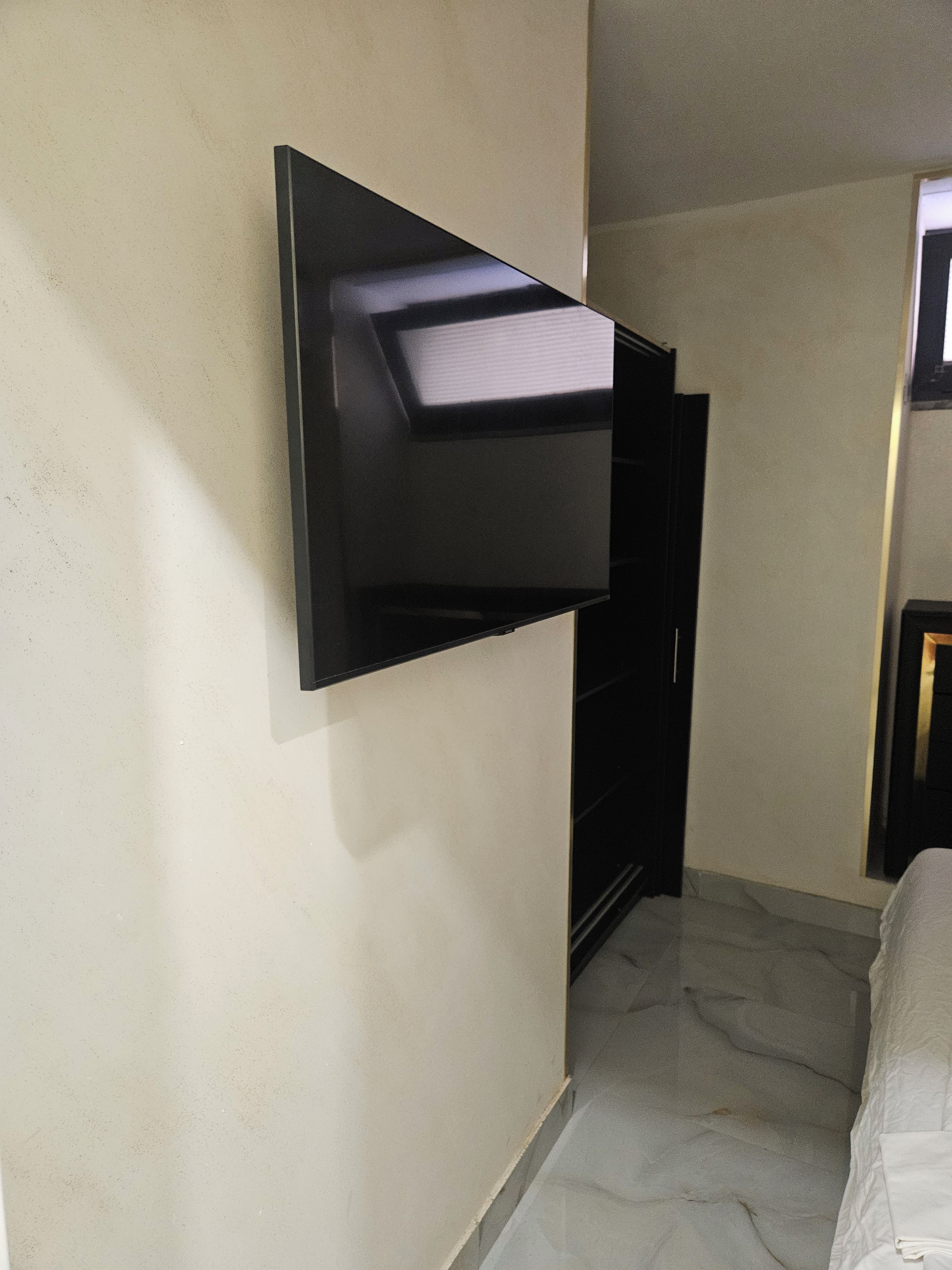 Une pièce avec un téléviseur fixé au mur, un lit avec une literie blanche et un sol en marbre.
