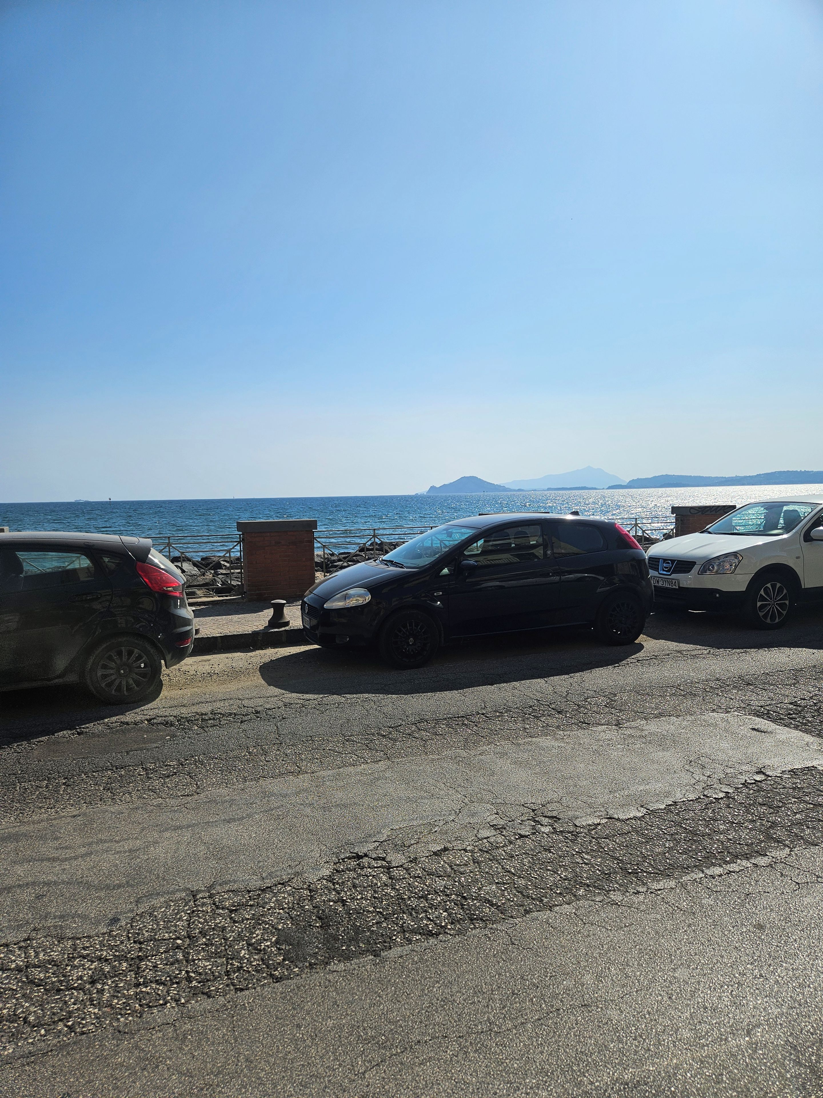 A parking area with vehicles, a clear sky, and a distant view of the sea and mountains.