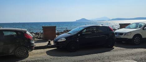 A parking area with vehicles, a clear sky, and a distant view of the sea and mountains.