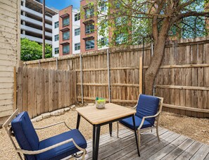 Eine Terrasse im Freien mit einem Holztisch und zwei blauen Stühlen, einem Holzzaun und einem Baum.