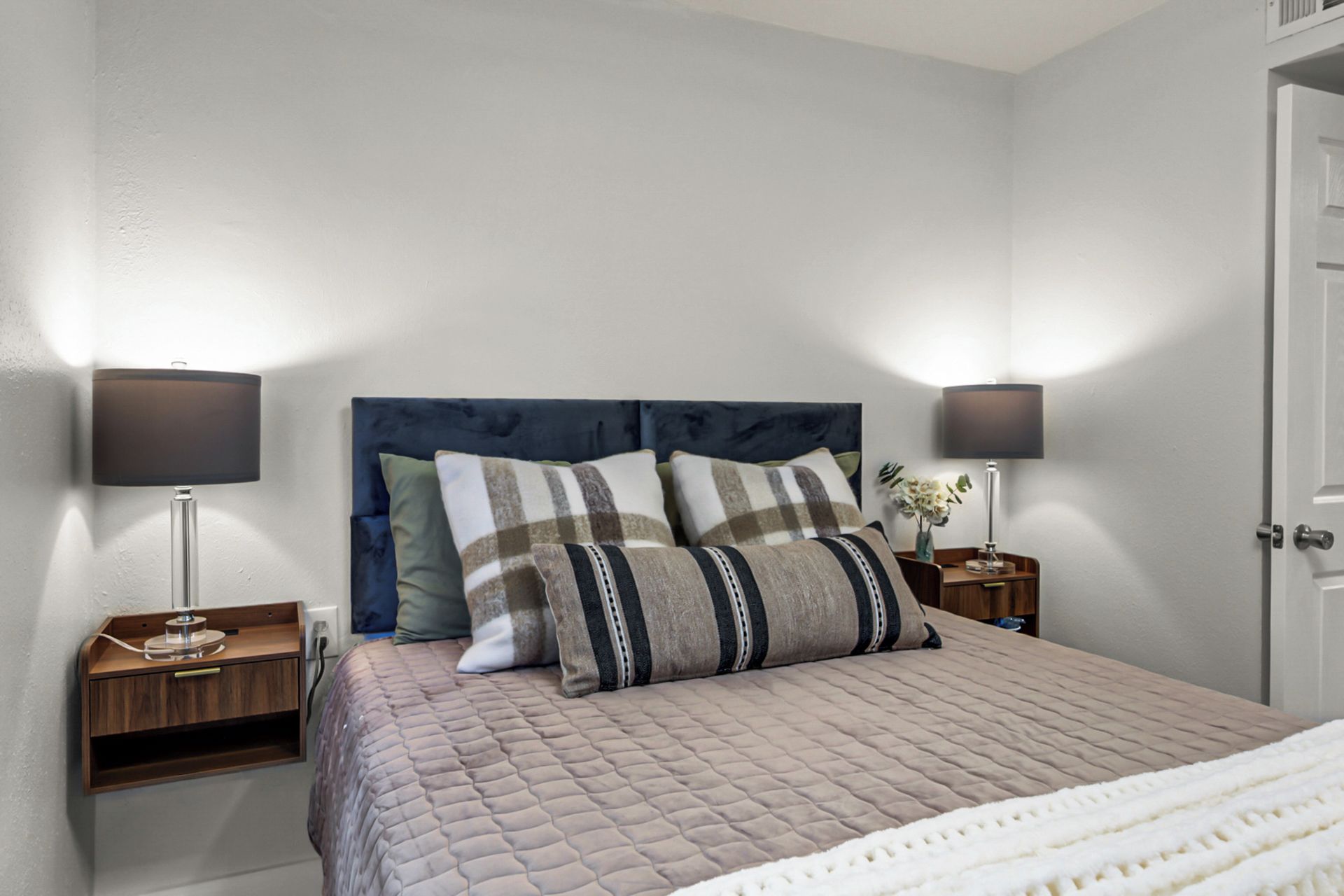 A neatly made bed with a quilted cover, two bedside tables with lamps, and a door to the right.