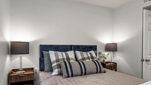 A neatly made bed with a quilted cover, two bedside tables with lamps, and a door to the right. - Northgate: Closest Home to Aggieland! 3BR, 2.5BA (College Station)