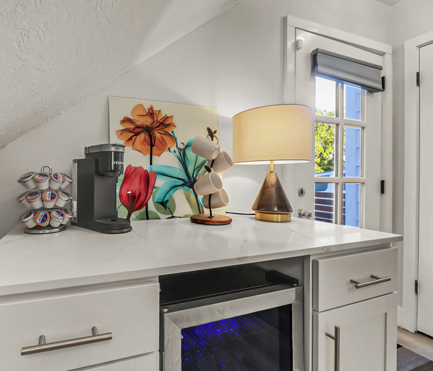 A kitchen counter with a coffee maker, a decorative flower painting, a lamp, and a window with blinds.