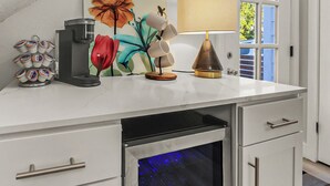 A kitchen counter with a coffee maker, a decorative flower painting, a lamp, and a window with blinds. - Northgate: Closest Home to Aggieland! 3BR, 2.5BA (College Station)