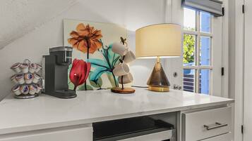 A kitchen counter with a coffee maker, a decorative flower painting, a lamp, and a window with blinds.