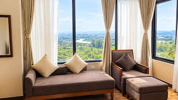 A room with a sofa and ottoman, large windows with curtains, and a view of a cityscape.