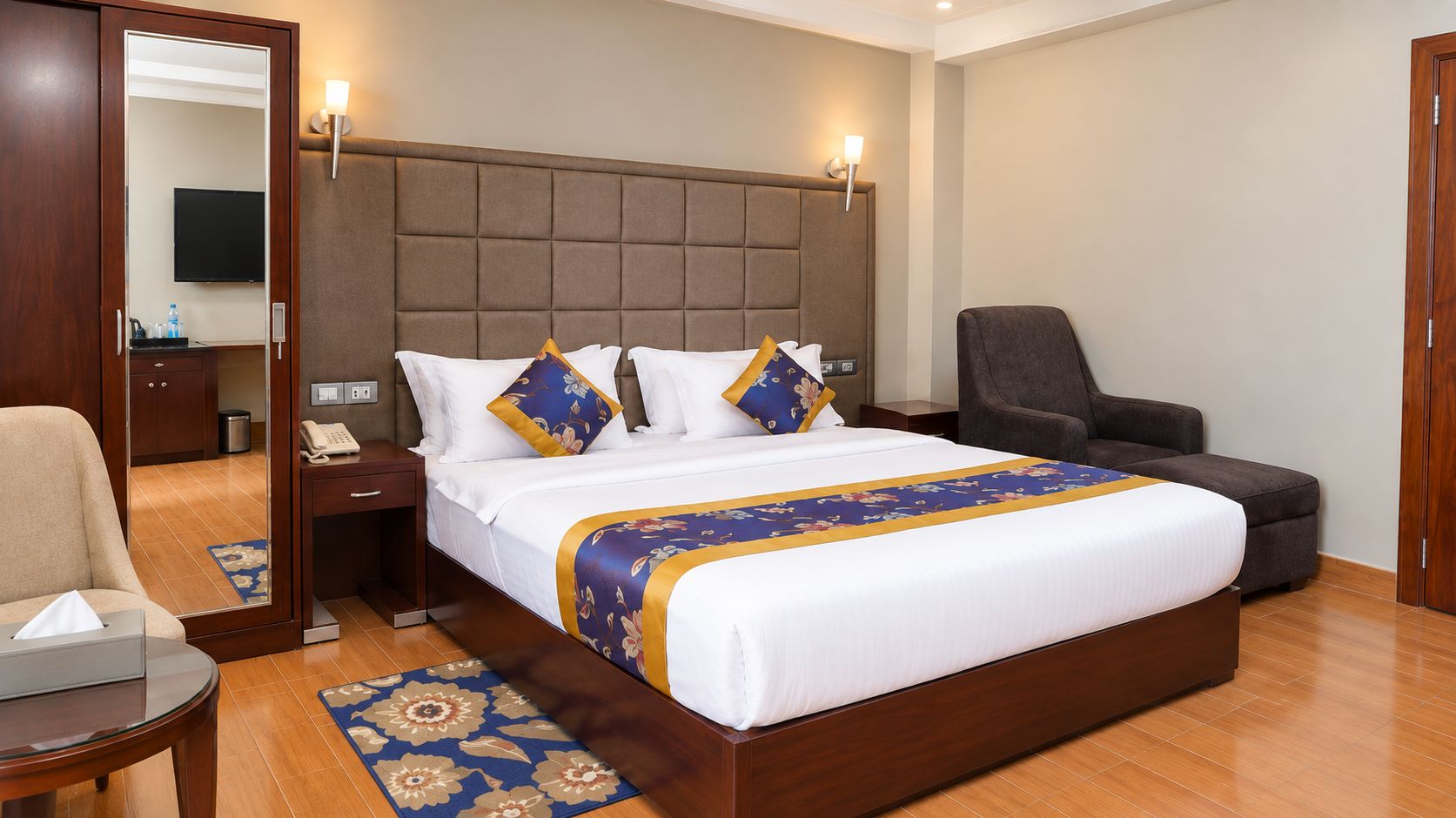A hotel room with a large bed, a bedside table with a lamp, a chair, a desk with a telephone, and a mirror.