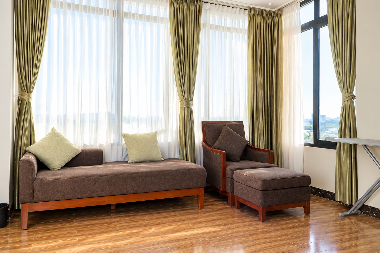 A room with a sofa, armchair, and ottoman, a window with curtains, and a view of the outside.