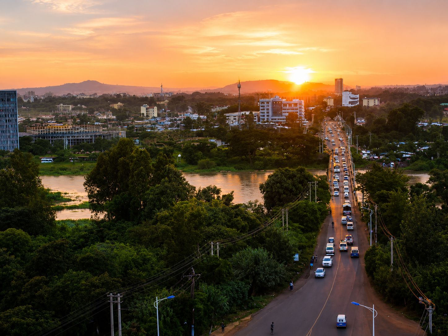 Une vue urbaine avec une rivière, des bâtiments et une route où circulent des véhicules, le tout baigné par les couleurs du coucher de soleil.