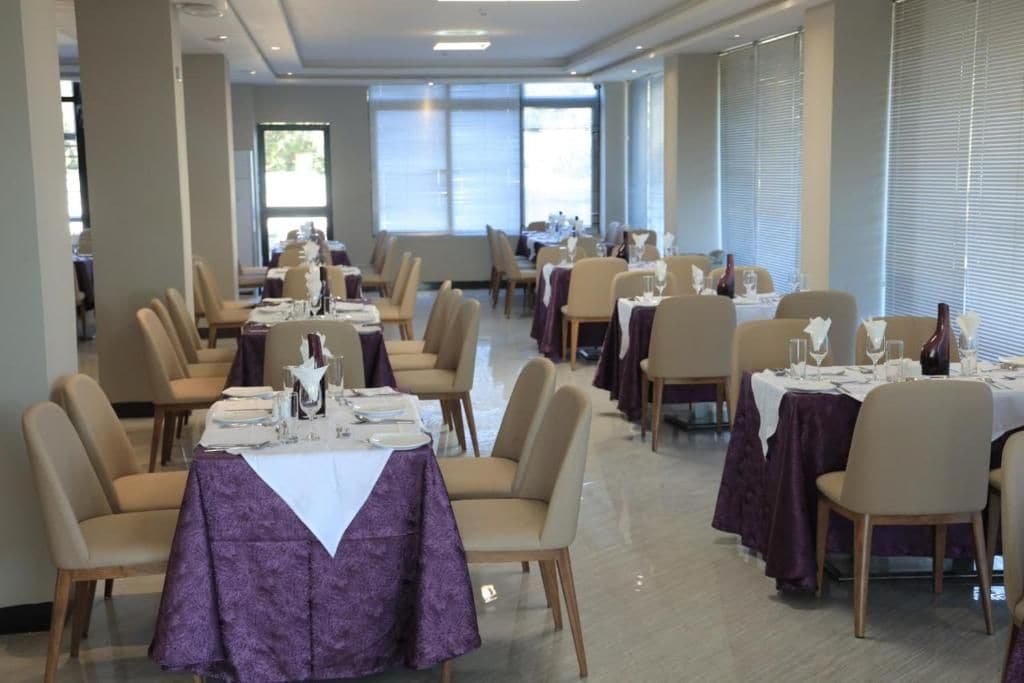 Uma área de refeições com várias mesas preparadas para o serviço, cadeiras dispostas ao redor de cada uma, e grandes janelas que oferecem luz natural.