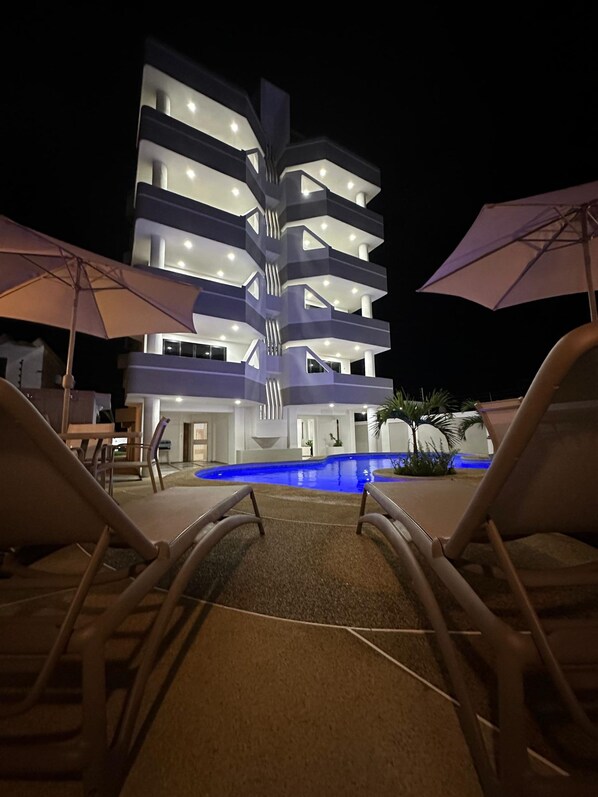 A multi-story hotel with a pool, lounge chairs, and umbrellas.