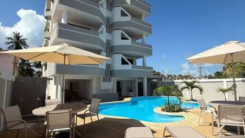 A modern building with multiple balconies, a swimming pool, and outdoor seating with umbrellas.