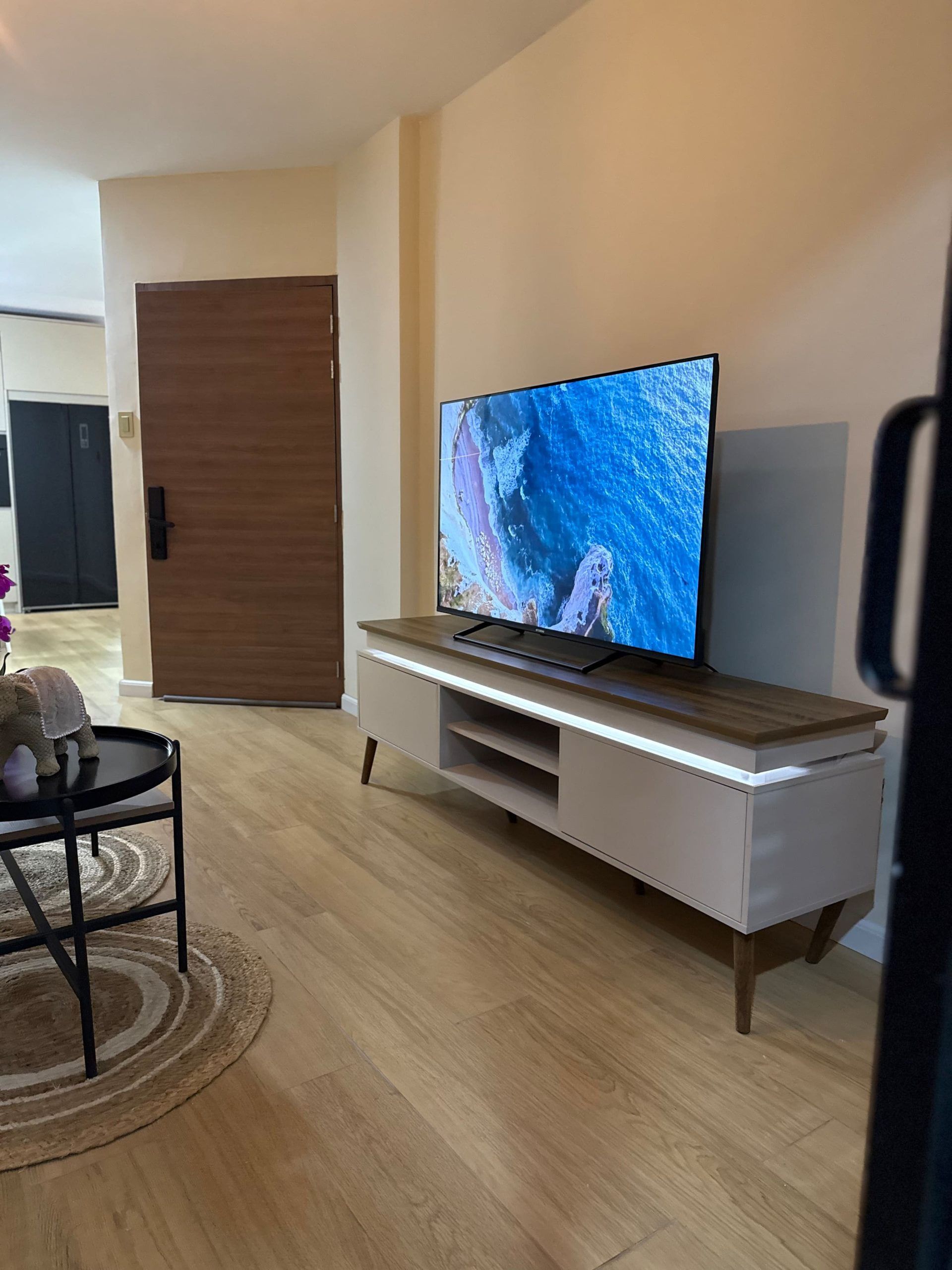 Ein modernes Wohnzimmer mit einem Flachbildfernseher auf einem Holzständer, einem kleinen runden Tisch mit einem Stuhl und einer Tür im Hintergrund.