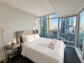 Ein modernes Hotelzimmer mit einem großen Bett, einem Nachttisch mit Lampe und Blick auf die Stadt durch raumhohe Fenster.