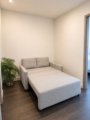 Quarto compacto com uma cama de solteiro, um vaso de plantas e piso de madeira.