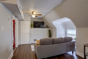A living room with a gray sofa, a ceiling fan, a television, and a window with blinds.