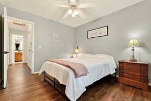 A bedroom with a bed, a nightstand, a dresser, a ceiling fan, and a hallway leading to a bathroom. - Wooded Whitetail Retreat at Beaver Lake - Sleeps 14 - Games, Decks & Lake Access (Rogers)