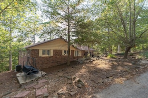 A single-story brick house with a sloped roof, surrounded by trees and a rocky ground. - Wooded Whitetail Retreat at Beaver Lake - Sleeps 14 - Games, Decks & Lake Access (Rogers)