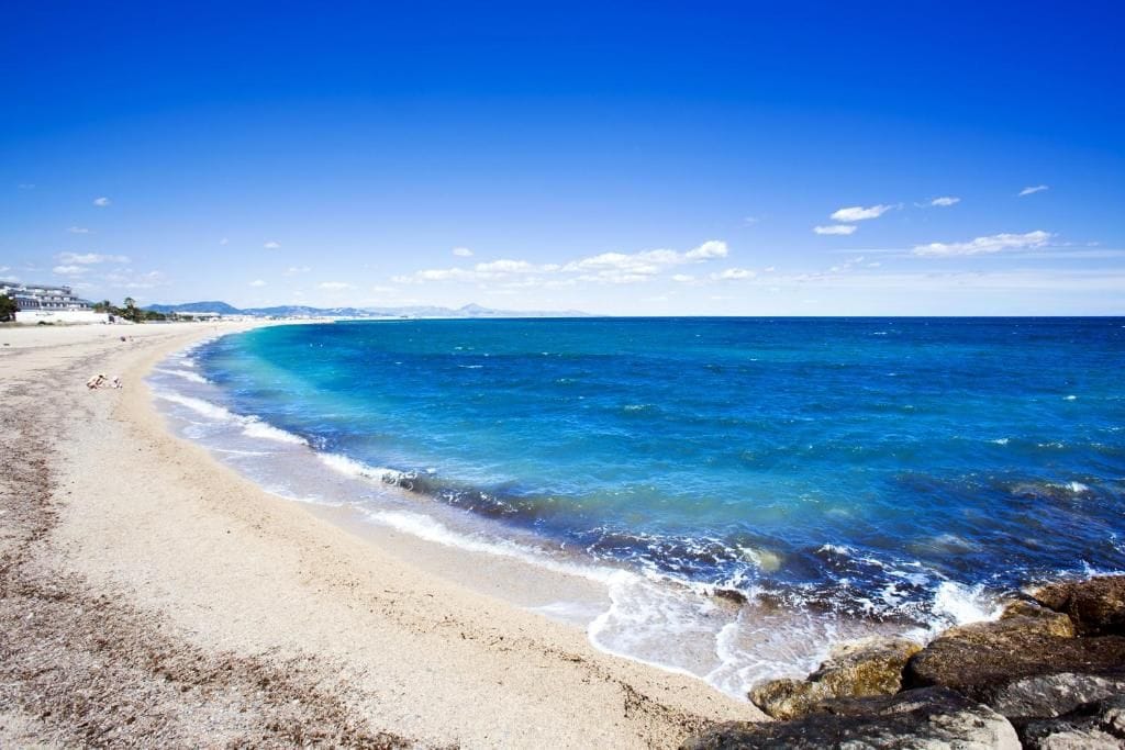 Ein Strand mit klarem, blauem Wasser, einem sandigen Ufer und einer entfernten Bergkette.